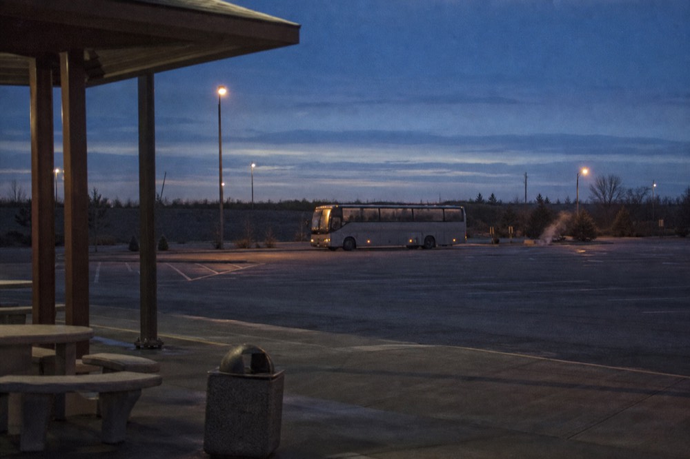 Highway service plaza at five in the morning with a single coach idling under sodium lights, empty parking bays, condensation on the windows, no logos visible