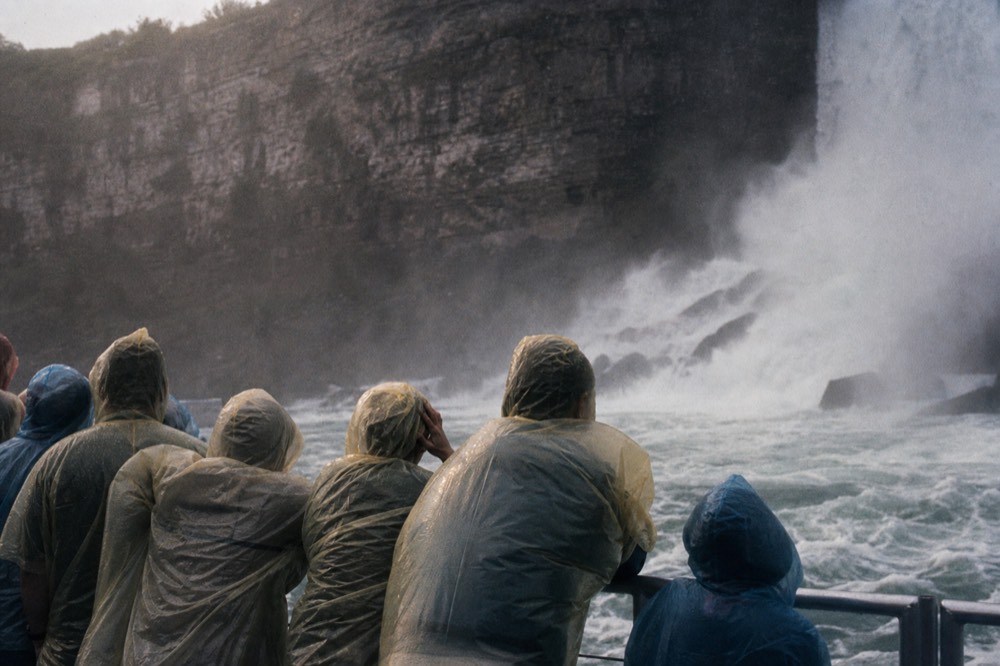 Hooded ponchos crowded at a boat rail from behind, heading into heavy waterfall spray, white water churning, sheer cliff face towering above in flat grey light