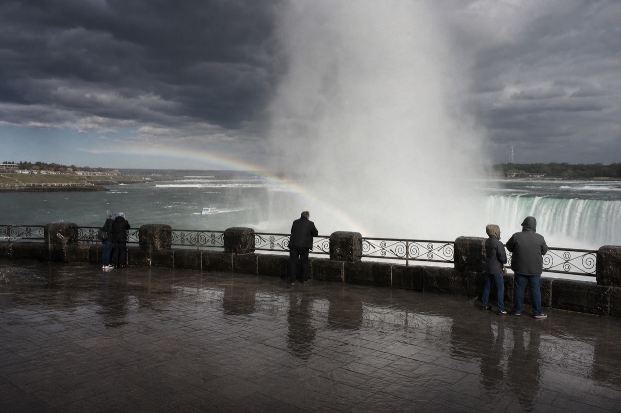Horseshoe-shaped waterfall seen from a wet observation deck at midday, thick vapour column rising, faint rainbow arc in the spray, dark soaked stone underfoot