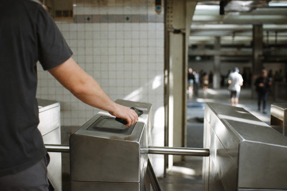 A row of stainless steel turnstiles seen from behind, one hand holding a phone flat above a contactless reader, tiled station wall soft and out of focus