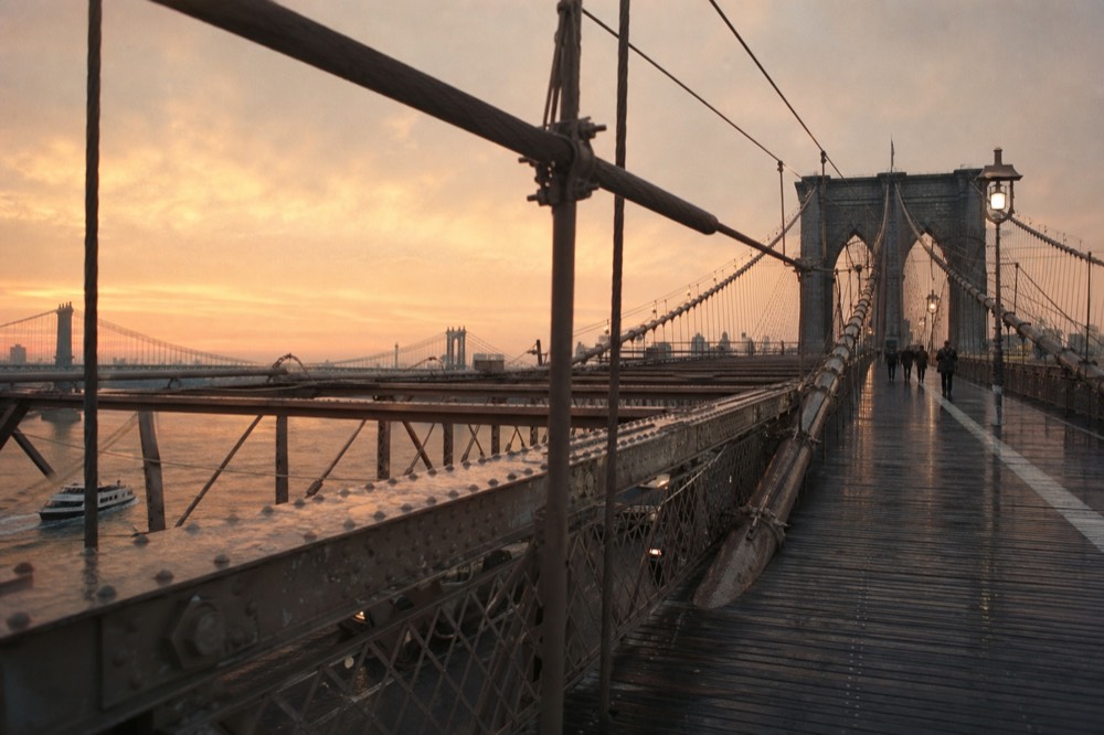 The Brooklyn Bridge walkway at sunrise seen from beneath the cables, weathered timber planks receding, pale granite arches against an empty apricot sky