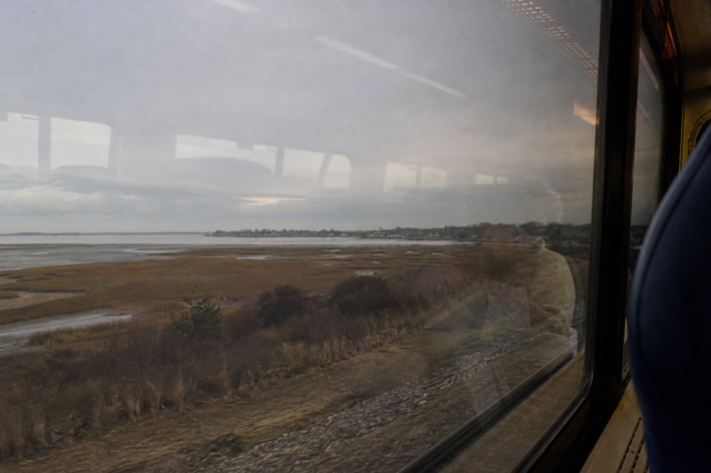 A train window view of coastal marsh grass and shallow water under flat grey light, the carriage interior faintly reflected in the glass