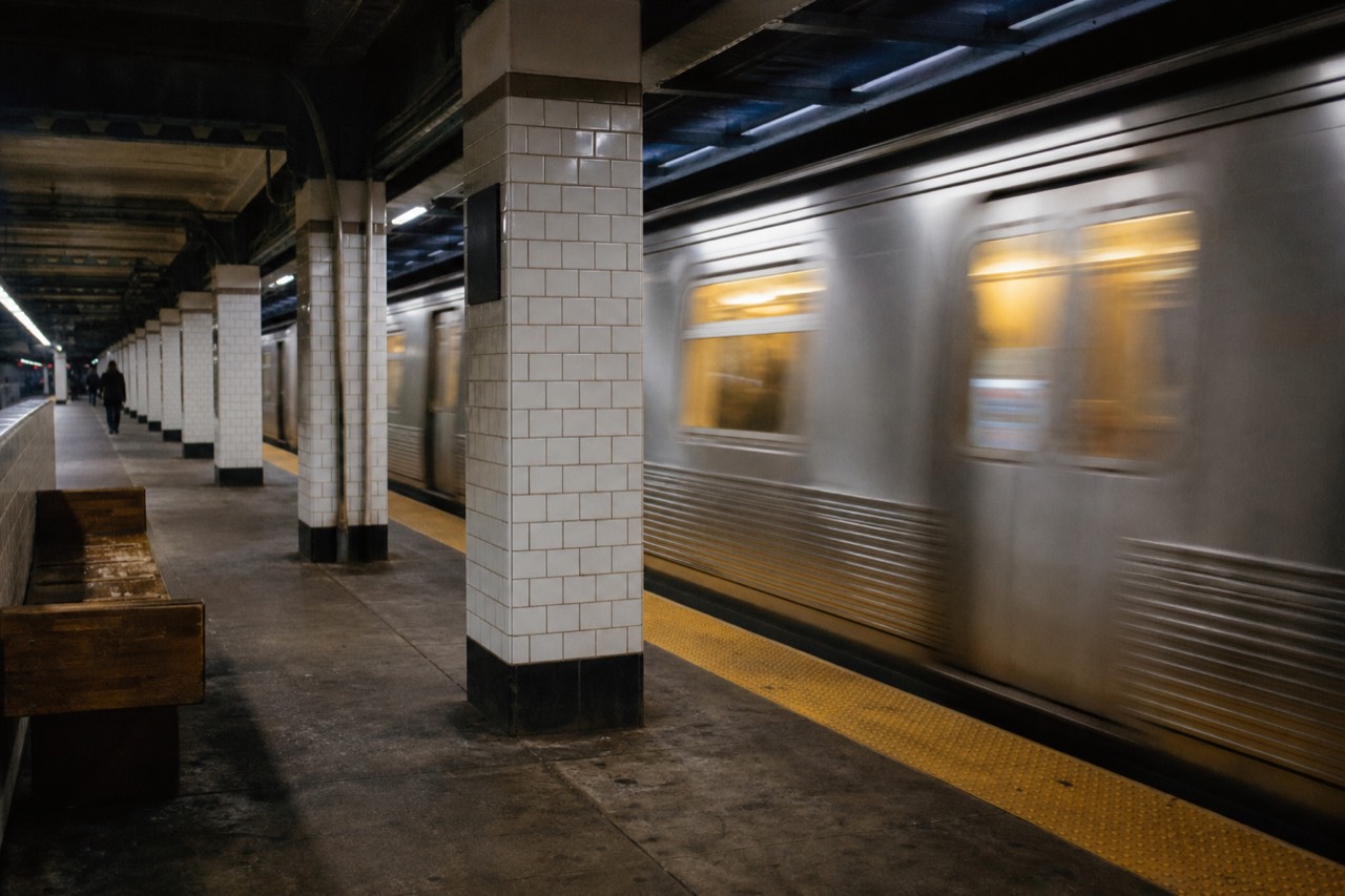 An underground subway platform at mid-morning, white tiled columns and painted steel beams, an empty wooden bench, warm light spilling from the windows of an arriving train