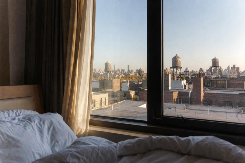 A quiet hotel room window at dawn with white bedding and half-open curtains, looking out on a grid of timber water towers and low brick rooftops