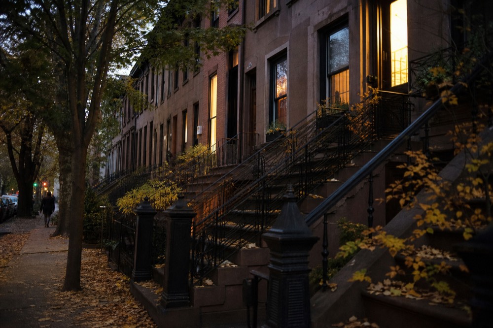 A row of brownstone stoops in autumn with fallen leaves drifted against the railings and warm lamplight showing in the parlor windows at dusk