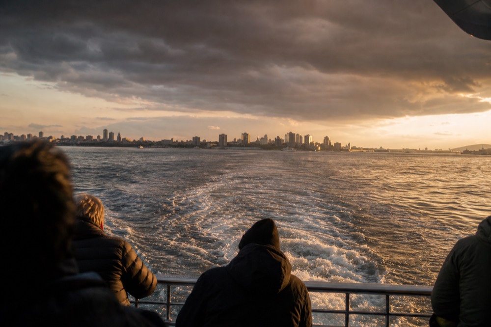Harbor water at golden hour from the open rear deck of a sightseeing boat, white wake churning behind, a low distant skyline on the horizon line