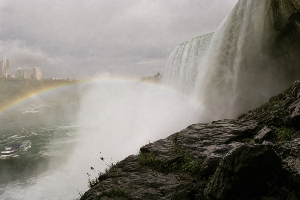 Thick white mist boiling up from the base of a wide waterfall, wet black rock in the foreground, a rainbow arcing through spray under flat grey sky