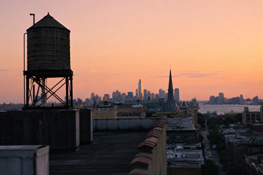 Empty rooftop water tower silhouetted against an orange dusk sky, tar-paper roofs stretching away, distant towers catching the last light