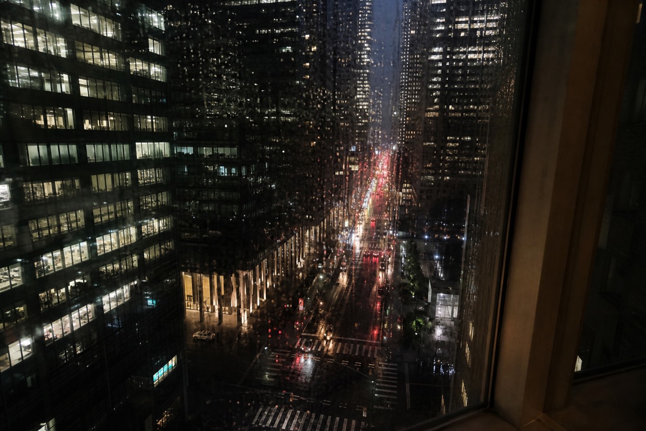A hotel window at night looking down a canyon of lit office floors, rain streaking the glass, traffic lights smeared red and gold on the wet street far below
