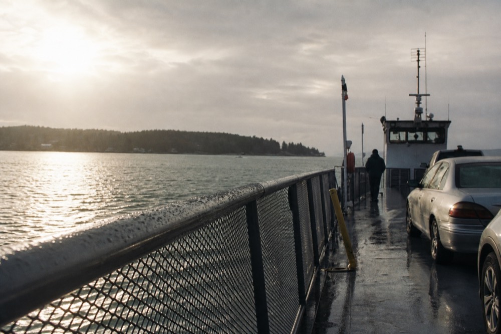 Low morning sun across a ferry deck's wet handrail, harbour chop grey-green below, a distant island shoreline rimmed with trees under thin cloud
