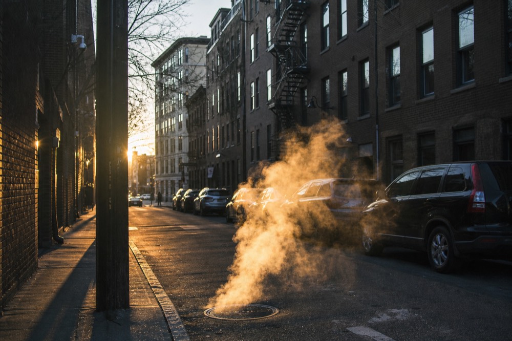 Steam rising from a manhole cover on a narrow downtown street at dusk, brick facades on both sides, a fire escape zigzagging up through cold blue light