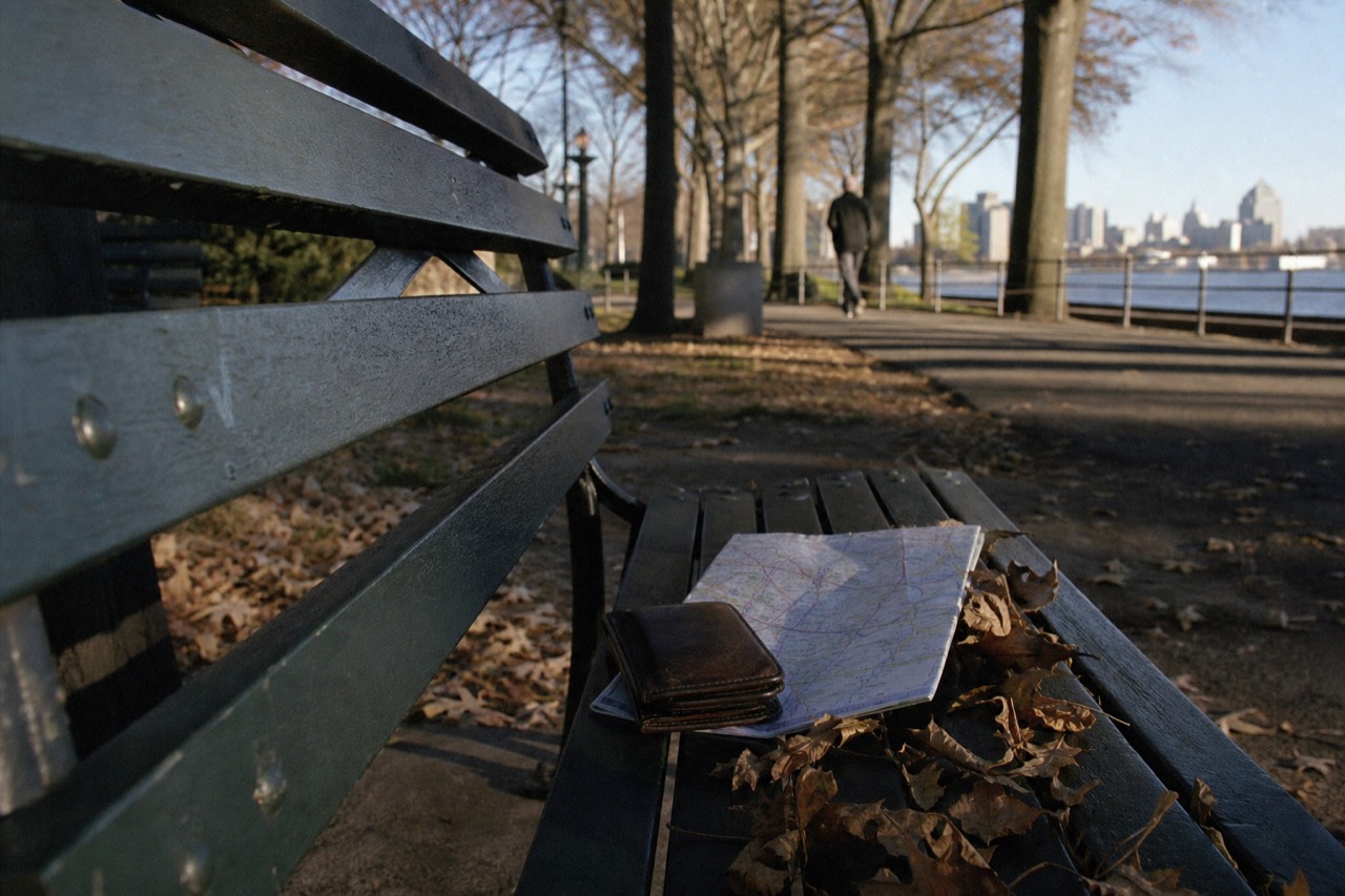 A worn leather wallet and a folded subway map on a park bench slat, autumn light through bare branches, dry leaves gathered along the seat edge