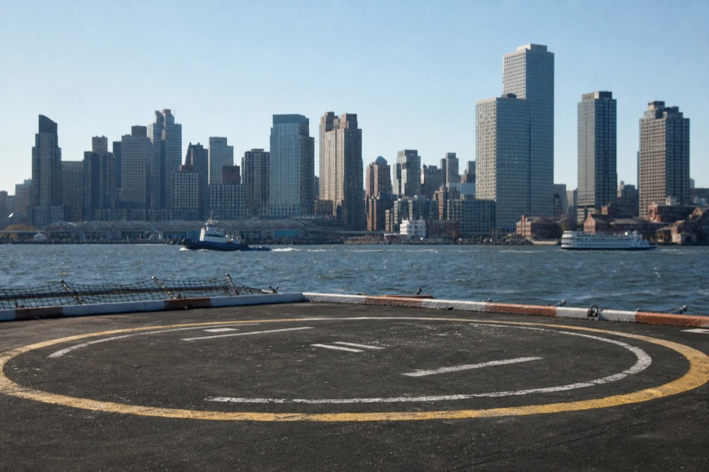 Empty helipad on a river pier at mid-morning, painted circle worn by rotor grit, harbor chop beyond the safety netting and a low grey skyline behind