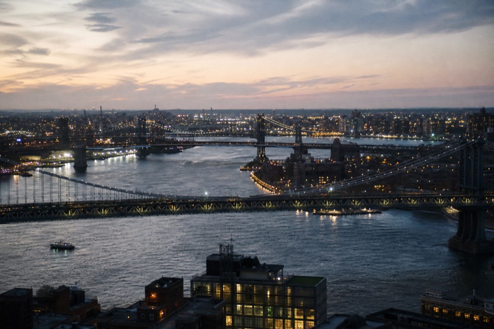 Looking down the East River from height at dusk, three bridges strung across dark water in sequence, Brooklyn lights spreading toward the horizon