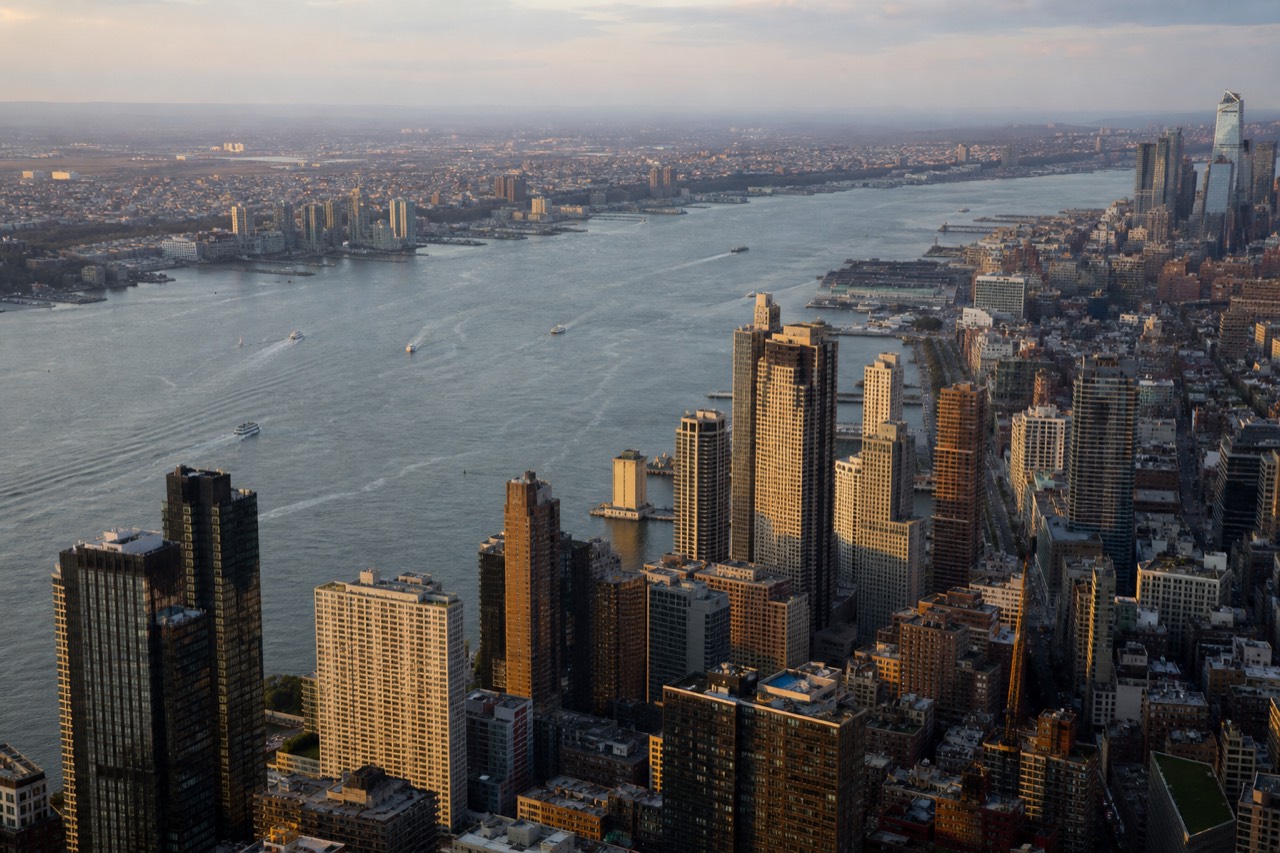Aerial view over the Hudson in clear late-morning light, Manhattan's towers packed edge to edge below, river traffic leaving thin white wakes on grey water