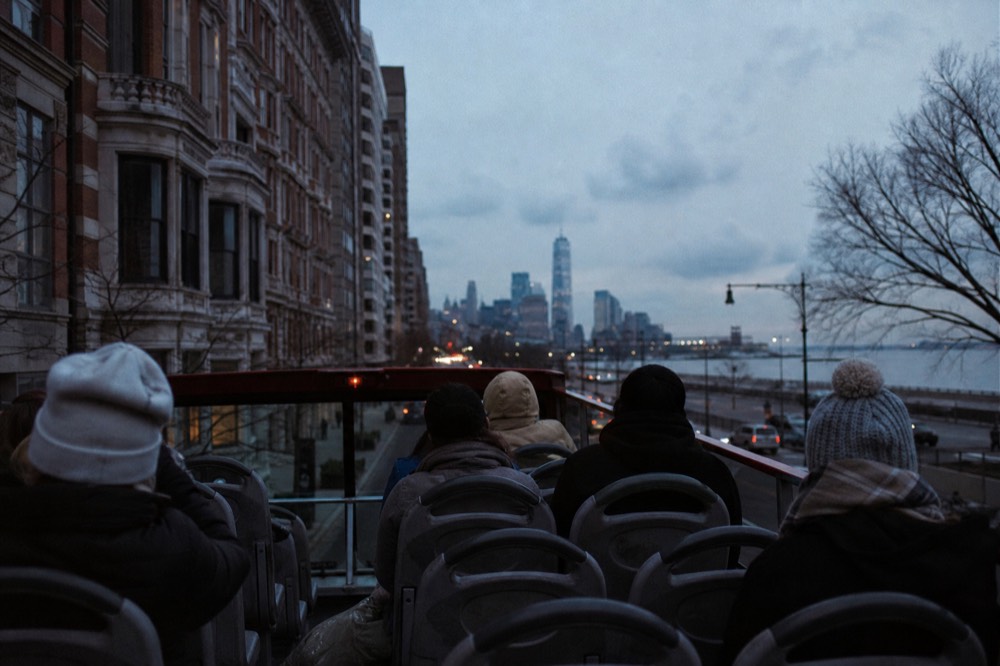 Passengers seated on the top deck of a stationary tour bus in slow avenue traffic, jackets zipped against wind, brownstone cornices passing at eye level