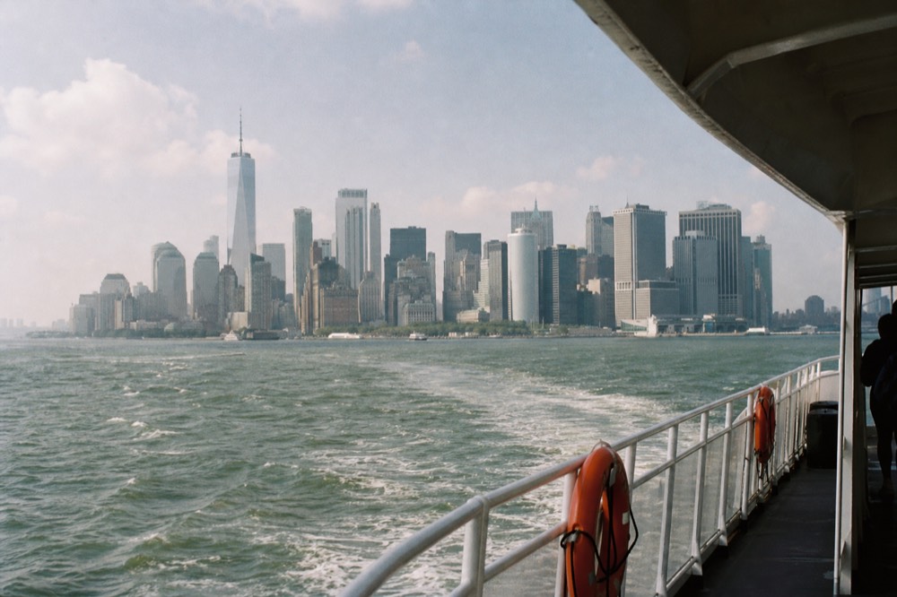 Ferry wake spreading across green harbor water at midday, Lower Manhattan towers rising behind a low haze, orange life rings mounted on a white rail