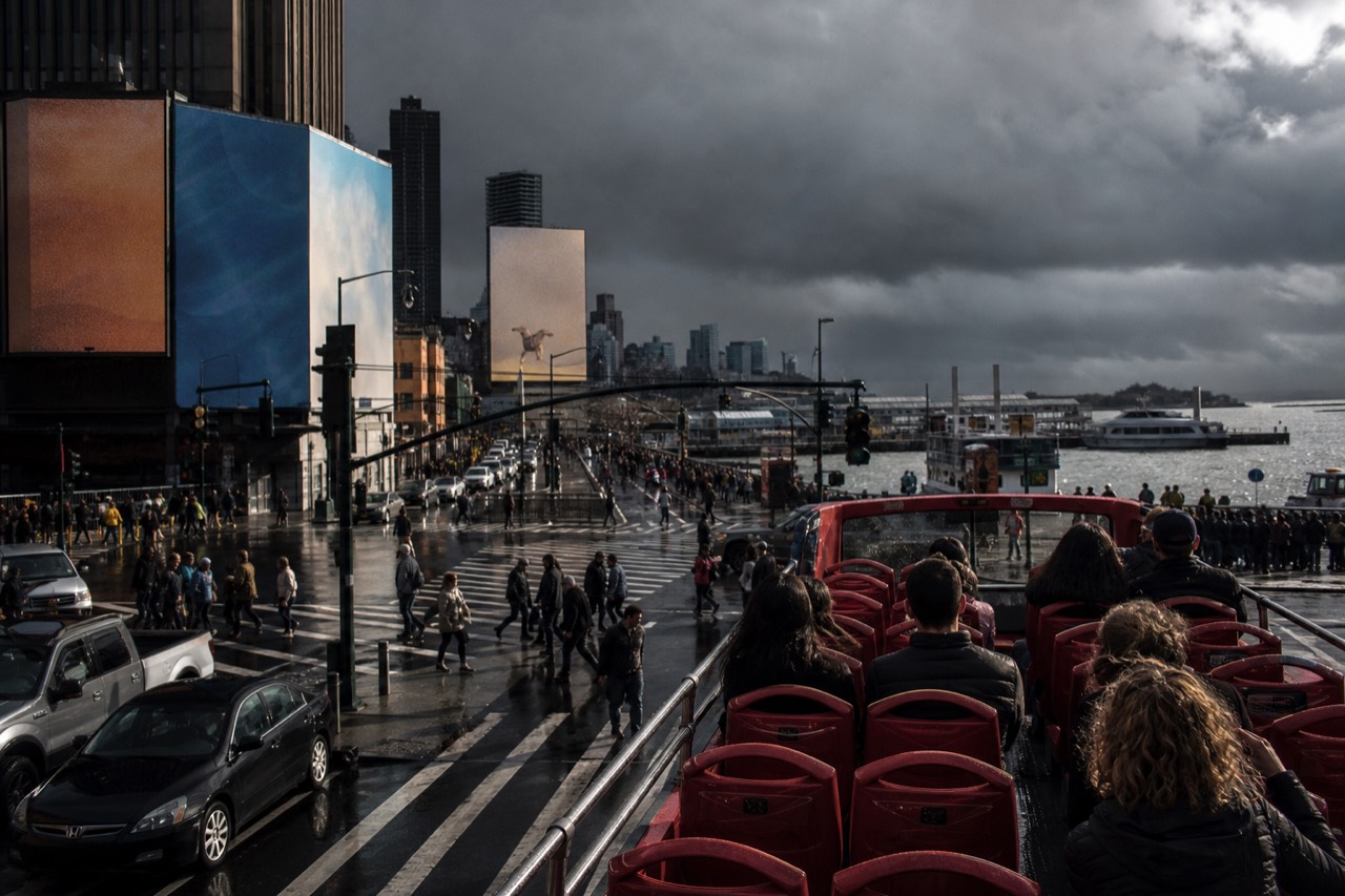Red open-top tour bus stopped at a crowded crosswalk under tall billboards, upper deck seats glossy with rain, grey afternoon light on wet asphalt