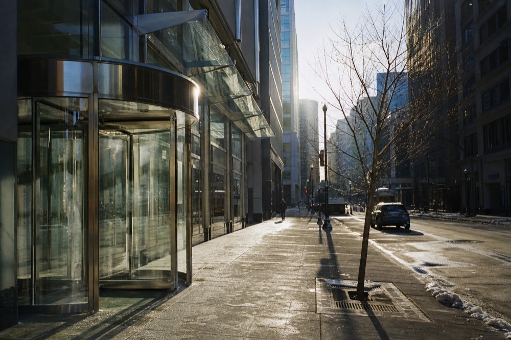 A steel and glass hotel facade on a cold clear morning, revolving door catching low sun, salt-stained sidewalk and a bare street tree in a metal grate