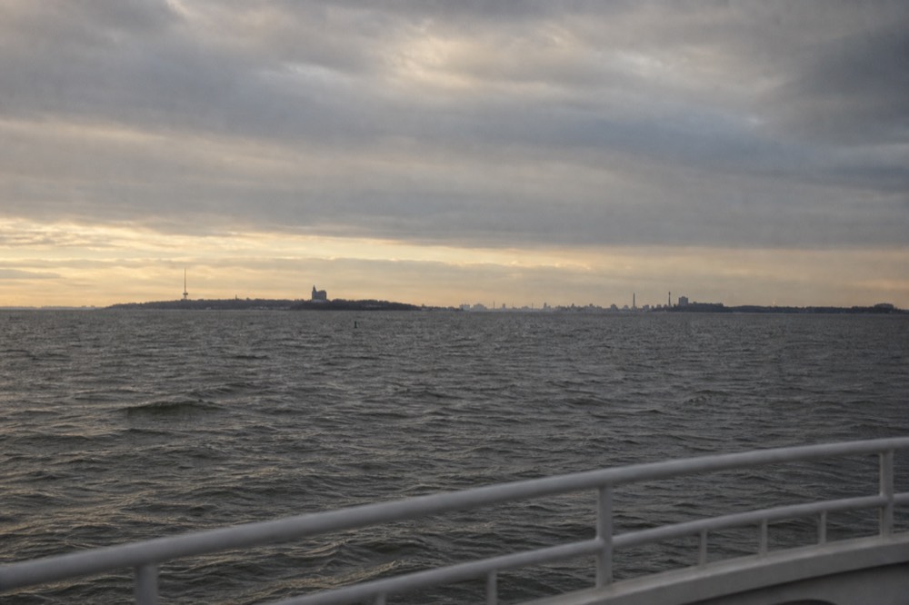 Grey harbour water under a flat winter sky, a bare boat rail in the foreground, distant island shoreline and low towers softened by cold sea haze