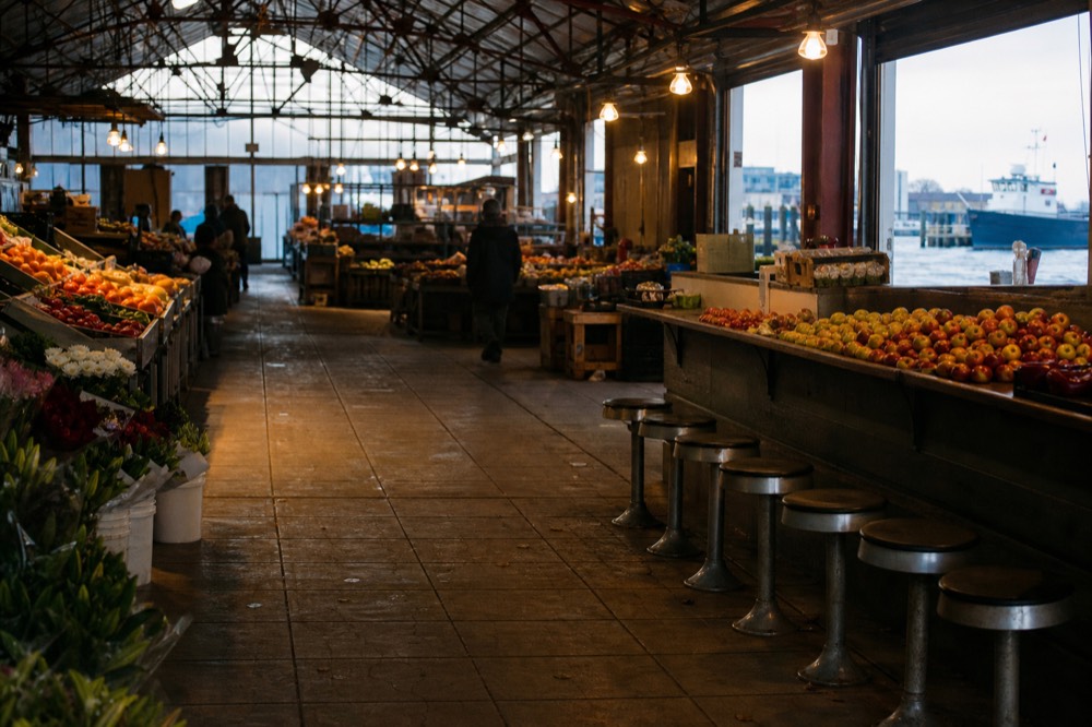 Busy indoor market hall with steel roof trusses and hanging bulbs, produce crates and counter stools, warm light falling on a worn tiled floor