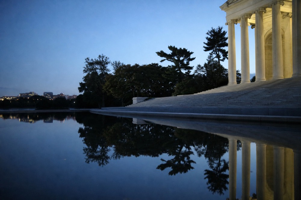 Marble memorial steps and tall white columns reflected in a long shallow pool at dusk, water perfectly still, deep blue sky above, no people in frame