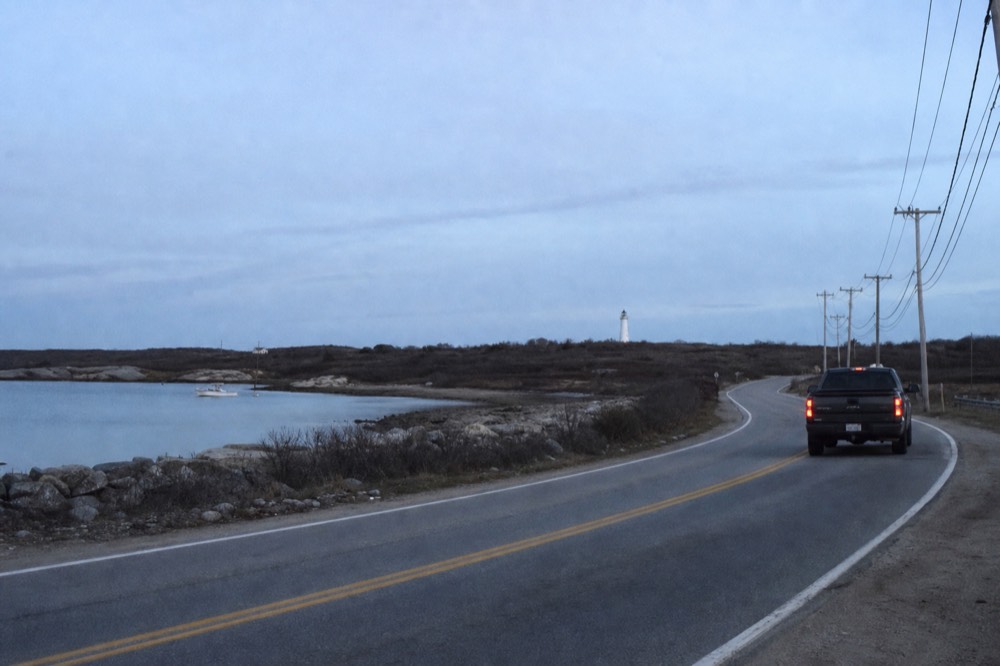 A two-lane road curving past salt marsh and low scrub under a wide pale sky, telephone poles receding, a white lighthouse tiny on the horizon.