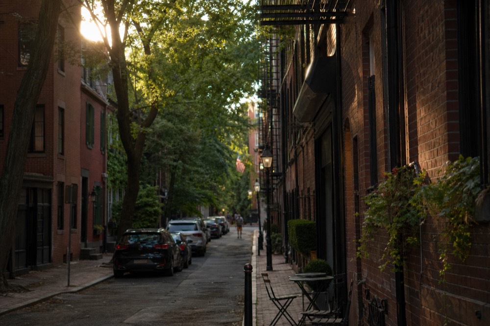 A narrow village street of low brick buildings and iron fire escapes, cafe tables on the sidewalk, dappled light through street trees overhead.