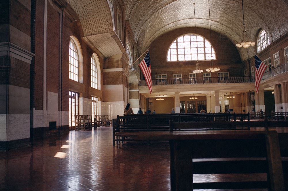 A red-brick immigration hall with a vaulted tiled ceiling and tall arched windows, dust in a shaft of afternoon light, empty wooden benches on a stone floor