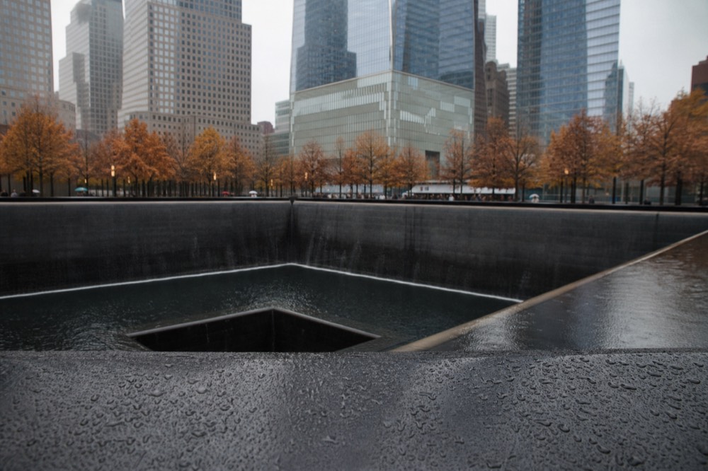 A square memorial pool of dark water falling over smooth stone edges, engraved bronze parapet in the foreground, glass towers rising blurred behind autumn leaves