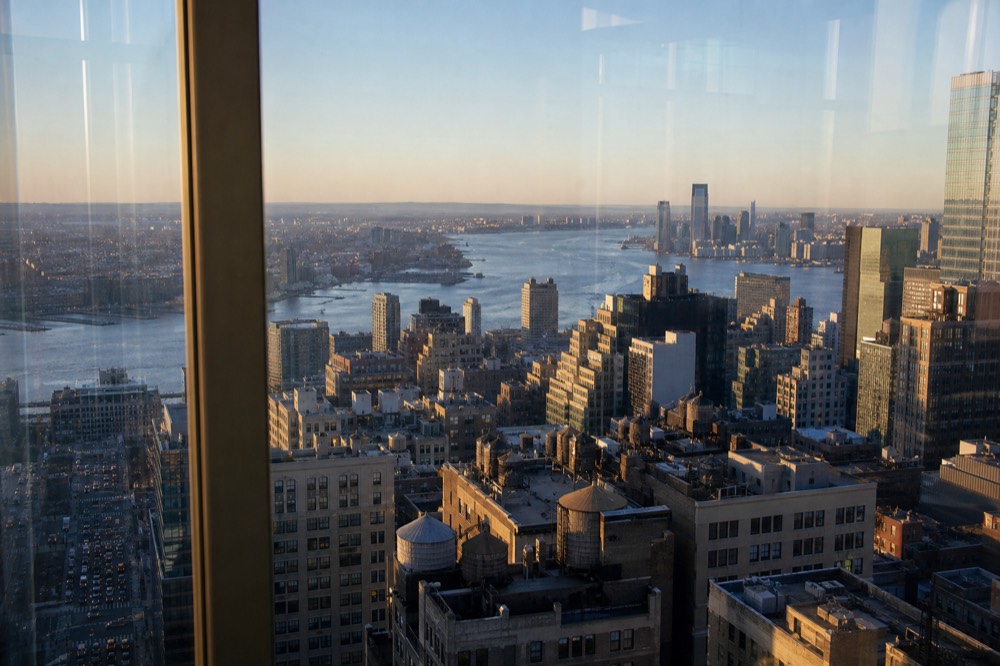 Midtown rooftops seen from a high glass deck at golden hour, water towers and setbacks casting long shadows, a river bending away toward flat distant land