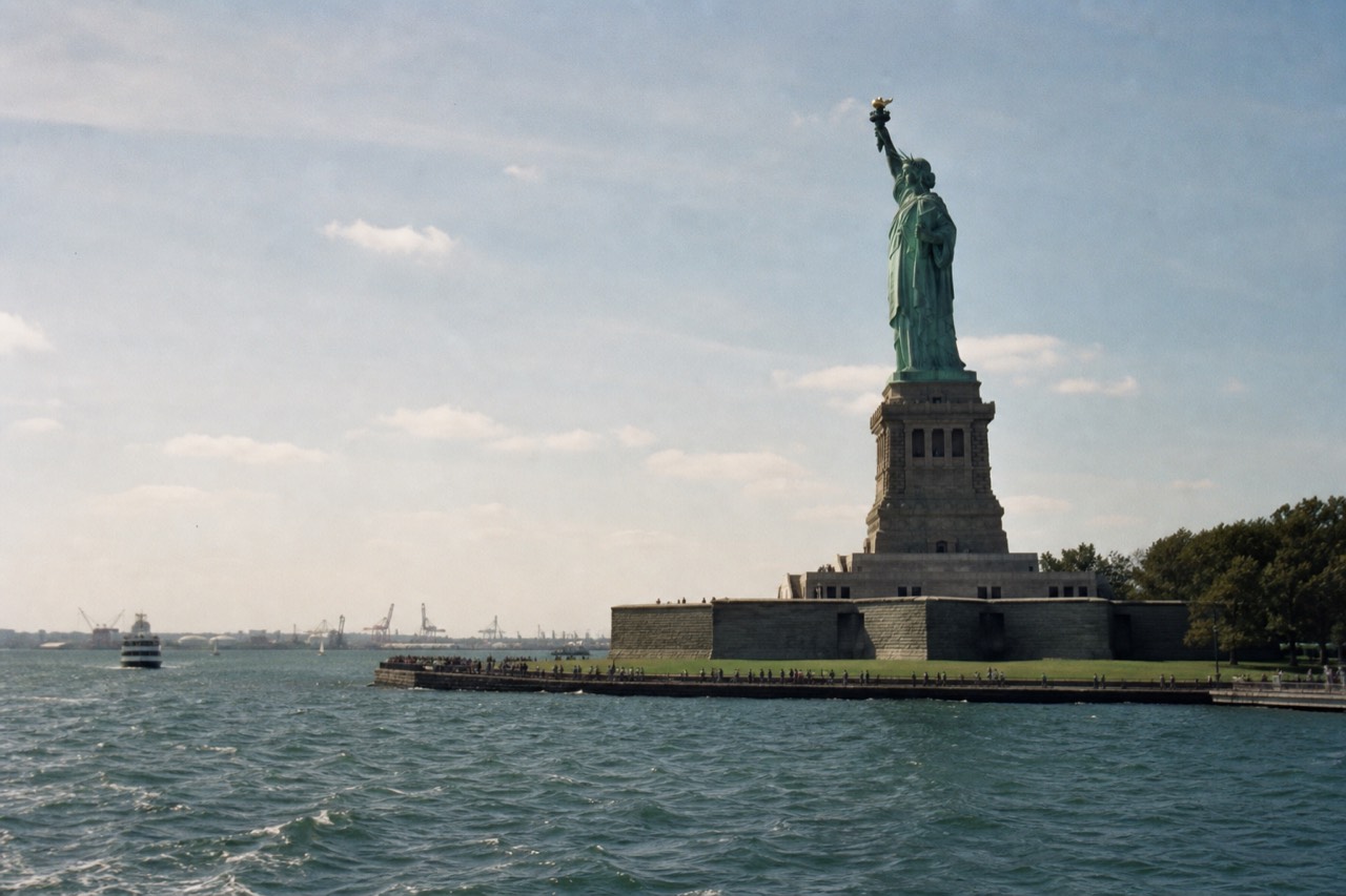 The green copper figure of a harbour statue seen from water level on a bright morning, gulls above the plinth, blue chop in the foreground and open sky behind