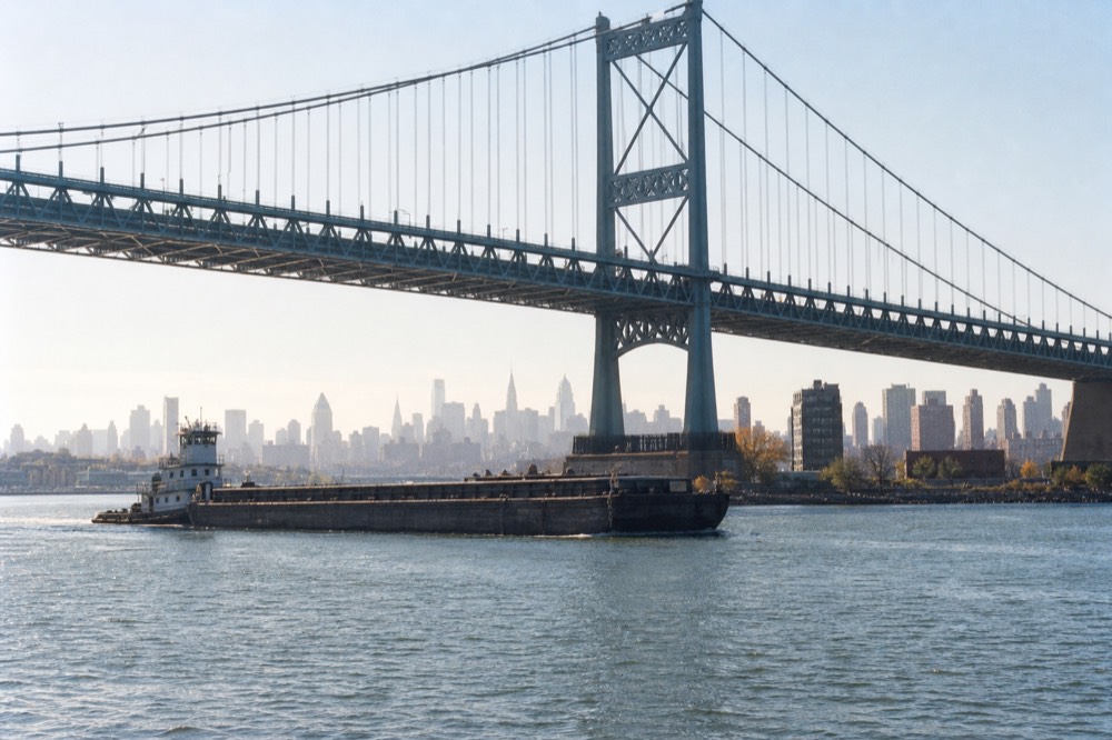 Steel suspension bridge photographed from water level, cables fanning above a passing barge, morning haze softening the far borough skyline behind