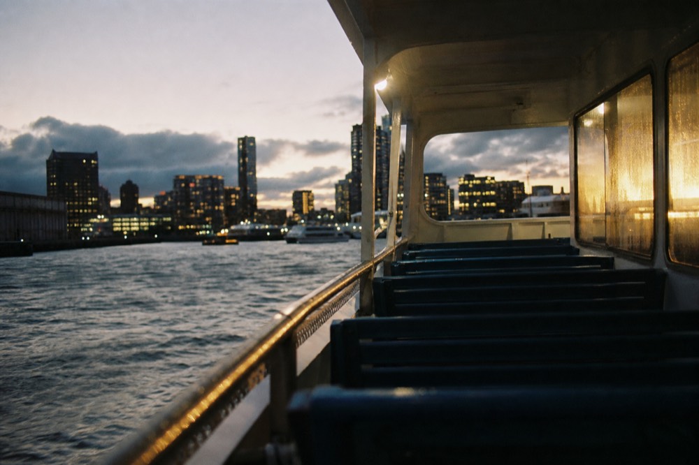 Empty passenger deck of a harbor boat at dusk, blue bench seats in rows, brass handrail catching light, city glow smearing across wet window glass