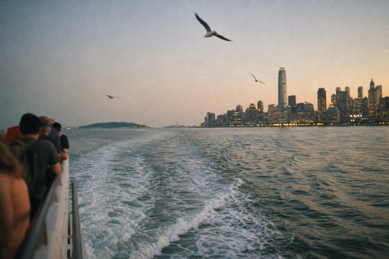 White wake spreading across green harbor water behind a sightseeing boat's rail at golden hour, low island shape on the horizon, gulls tracking overhead