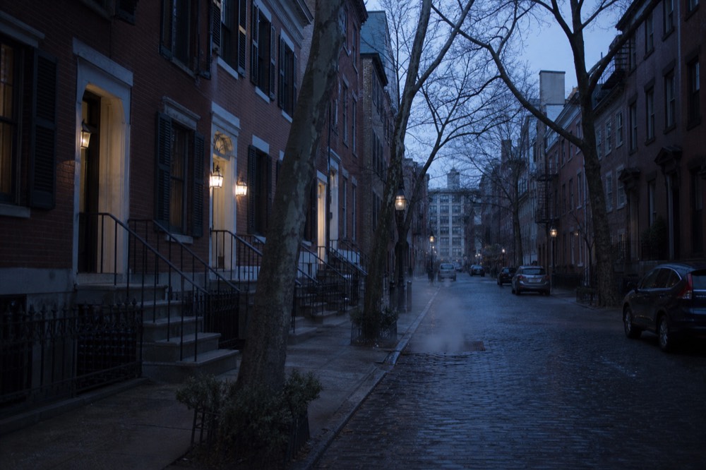 Narrow Greenwich Village street of low brick townhouses with iron stoops and bare plane trees, wet cobbles reflecting a pale winter sky, no shopfront signage