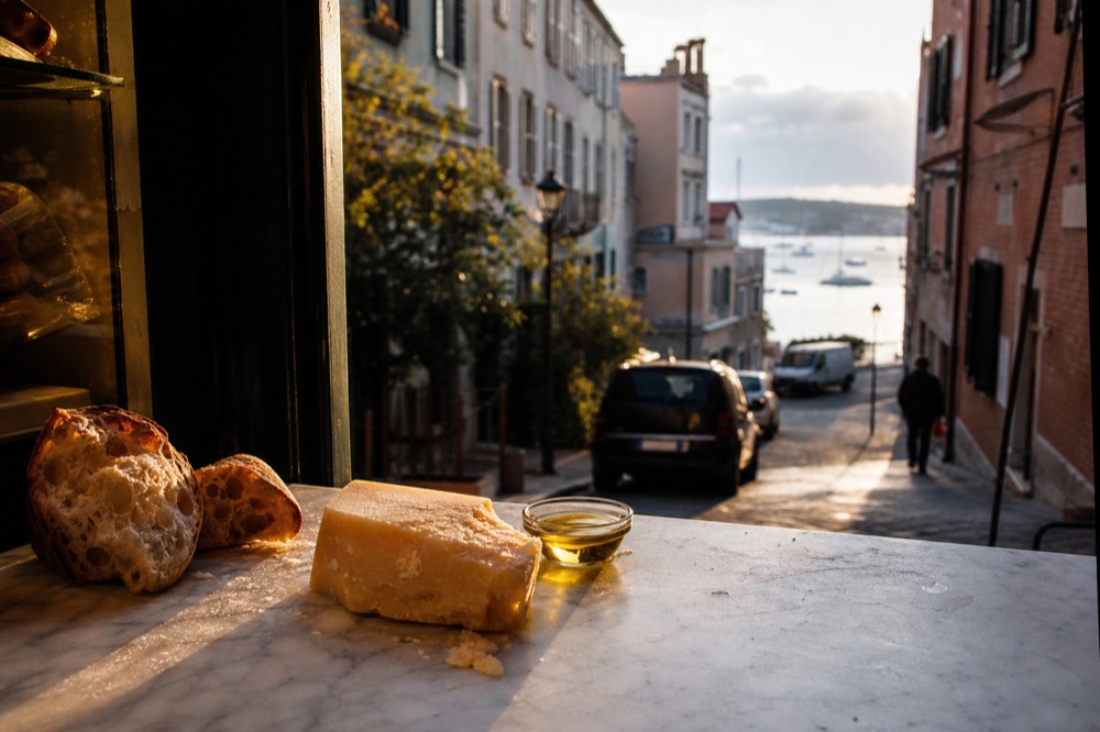 Marble counter dusted with flour holding torn bread, a wedge of hard cheese and olive oil in a small glass dish, warm shop light from the left