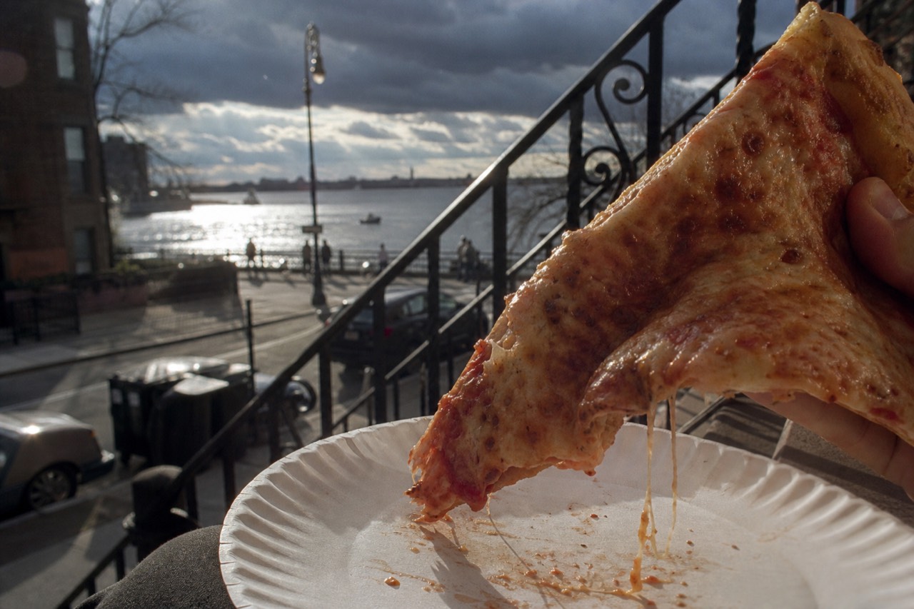 Close-up of a folded slice of pizza held over a paper plate on a sunlit brownstone stoop, cheese stretching, cast-iron railing blurred behind