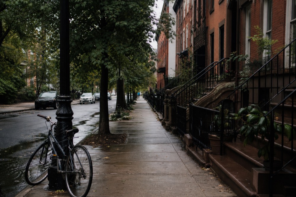 A narrow tree-lined street of red brownstone stoops, wrought-iron railings, late afternoon sun on brick, a bicycle chained to a lamppost