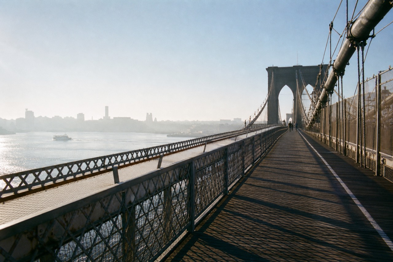 A wide steel suspension bridge walkway at dawn, cable shadows striping worn planks, mist over the water below, empty pathway curving toward stone towers
