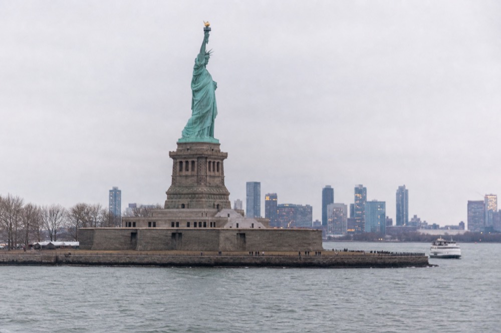 A copper-green statue seen from harbor level in flat morning light, pedestal stonework rising from the island, water and pale sky filling the rest of the frame