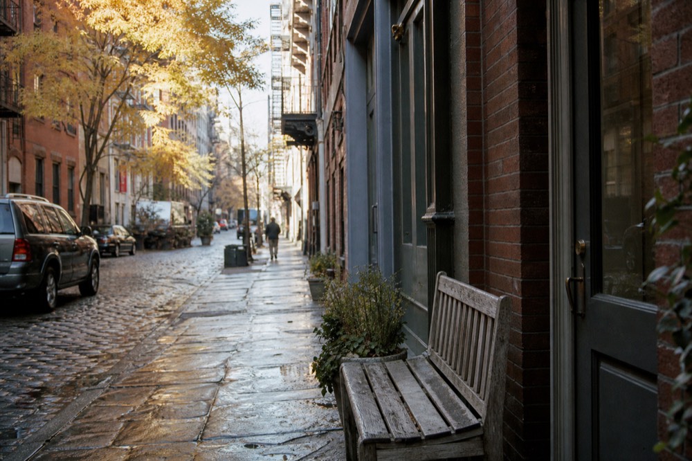 A narrow Greenwich Village side street in soft afternoon light, low brick facades, a worn wooden bench outside a shopfront, cobbles still wet from rain