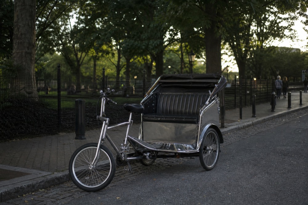 A pedicab parked at a park kerb in early evening, chrome frame and empty padded bench seat, plane trees and black iron fencing blurred behind