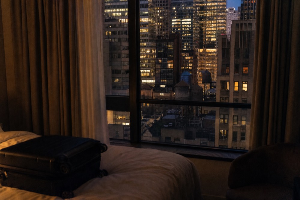 A hotel room window in Midtown at dusk, curtains half open, water towers and lit office floors stacked behind the glass, one suitcase unopened on the bed.