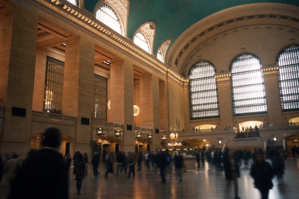 A grand railway concourse interior with a vaulted painted ceiling, shafts of daylight from tall arched windows, commuters reduced to soft motion blur below.