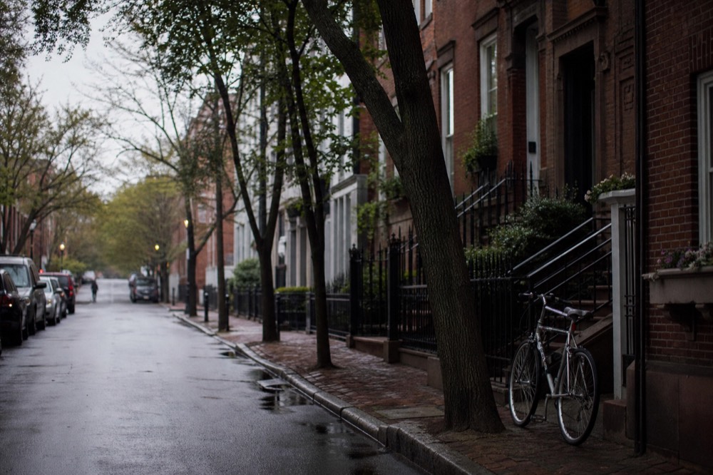A narrow tree-lined street of red brick townhouses with iron stoop railings, late afternoon shadows across the pavement, one bicycle chained to a railing.