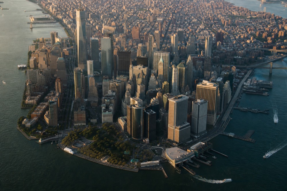 Aerial view of a dense island skyline seen from above the water at mid-afternoon, long building shadows, a river bend and two bridges visible at the frame edge.