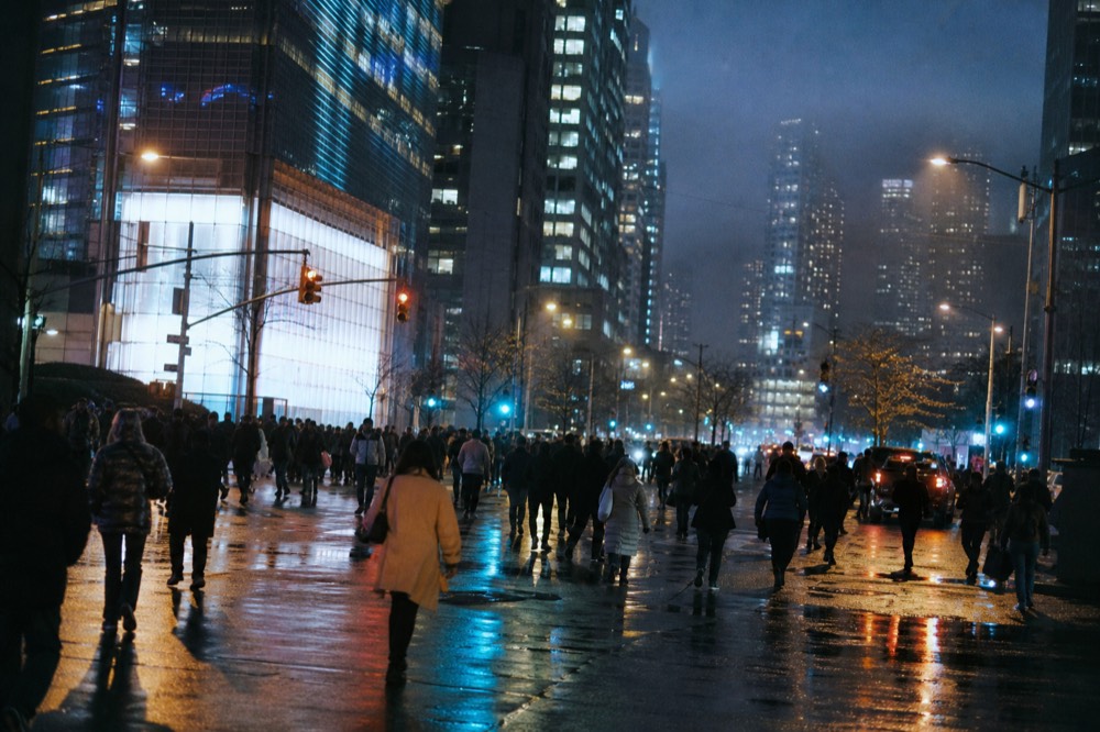 A crowded illuminated square at night, layered light from overhead screens washing across wet asphalt, silhouetted figures crossing in every direction.