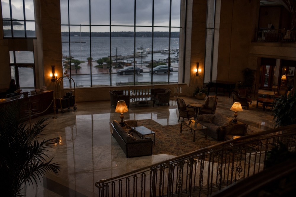 A hotel lobby seen from a mezzanine, warm lamplight on marble, luggage trolley at the desk, tall windows showing a grey street beyond.