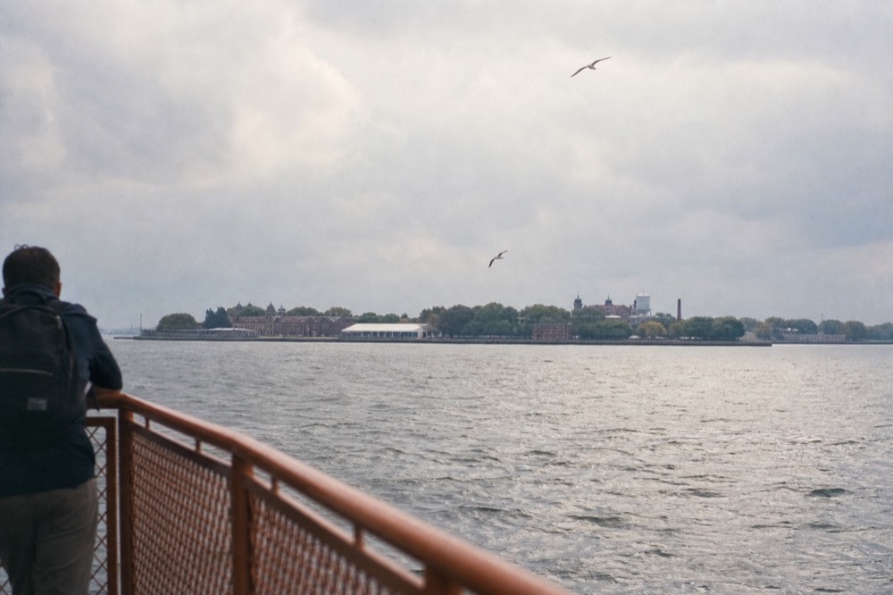 A ferry deck railing with harbor chop below, gulls angled against grey sky, a low green island and its brick museum buildings on the horizon.